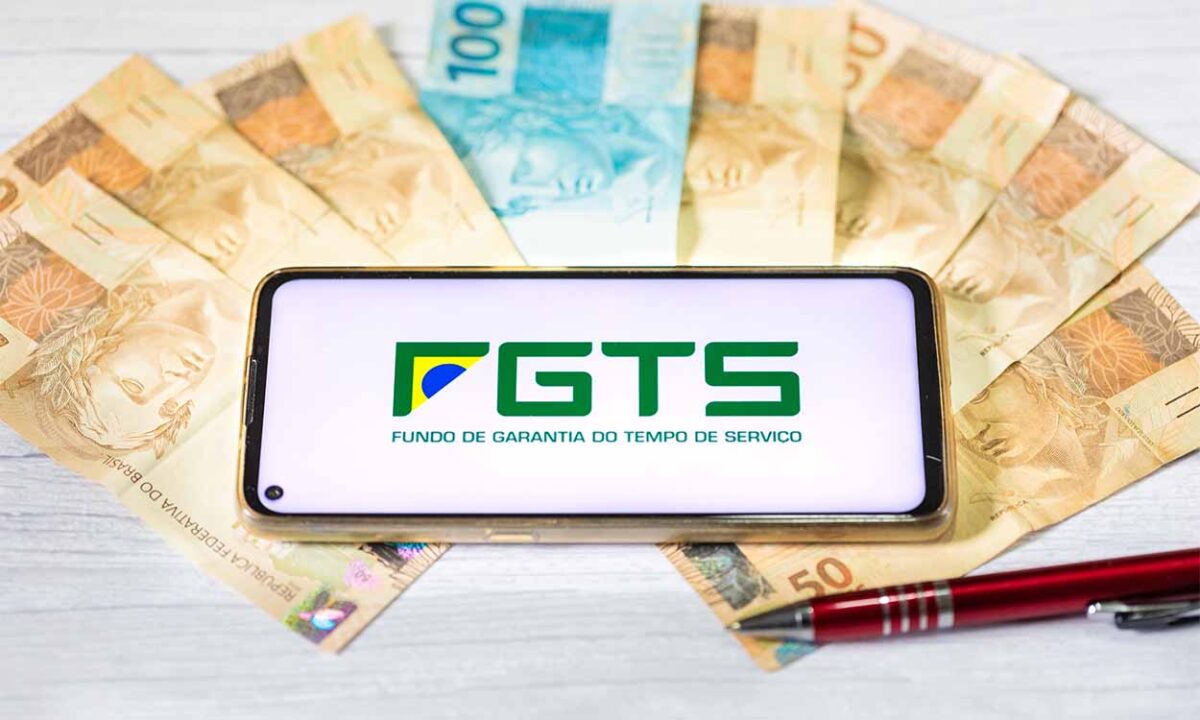 Leque de dinheiro, contendo notas de 50 e 100 reais, embaixo de um celular que mostra o logo do FGTS. Há, também, uma caneta em cima de uma nota