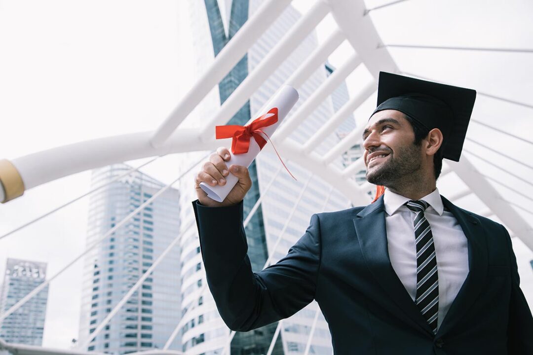 Homem usa capelo de formatura e apresenta diploma em mãos, próximo a uma escada e à paisagem urbana.