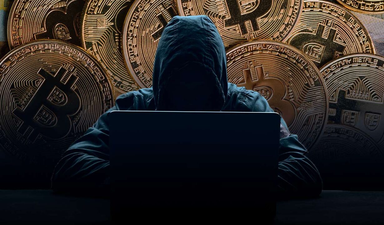 Hacker encapuzado atrás de um notebook, ao fundo tem várias criptomoedas de bitcoins.