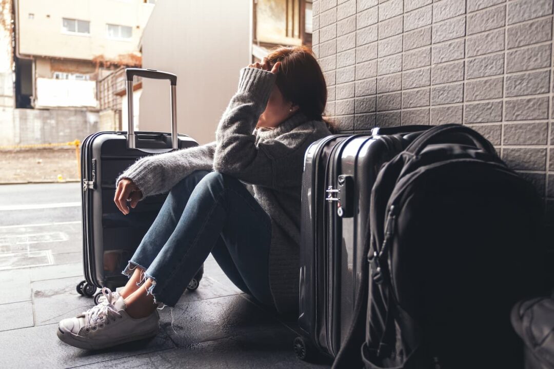 Na imagem, uma turista no aeroporto com as mãos na cabeça e fazendo expressão de tristeza, ao lado dela tem duas malas de viagem