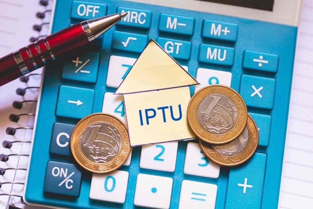 A sigla IPTU dentro de um papel em formato de casa, cercado de moedas, em cima de uma calculadora