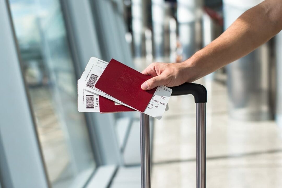 Mão de uma pessoa segurando passagens e a alça de uma mala de viagem, em aeroporto.