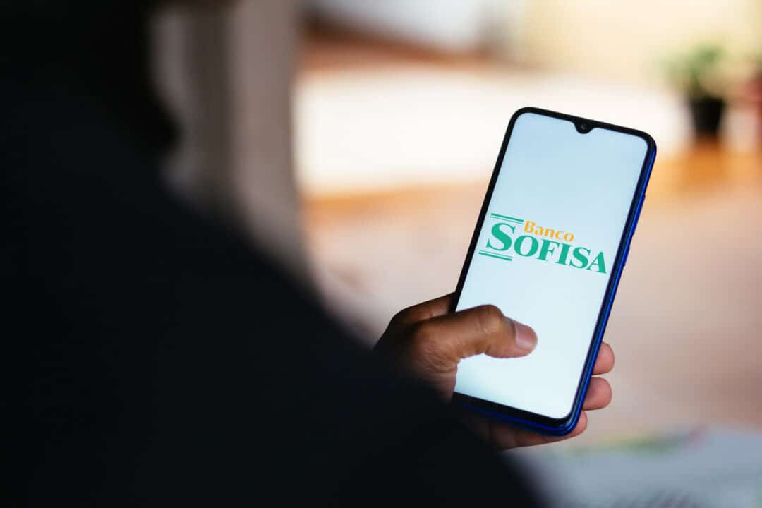 Pessoa de costas segurando celular que mostra logo do banco Sofisa Direto