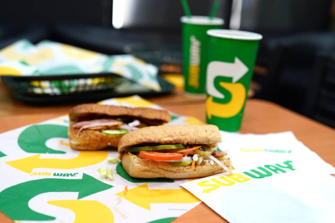 Dois sanduíches da Subway e um refrigerante estão dispostos na mesa, acompanhados por guardanapos personalizados da marca.