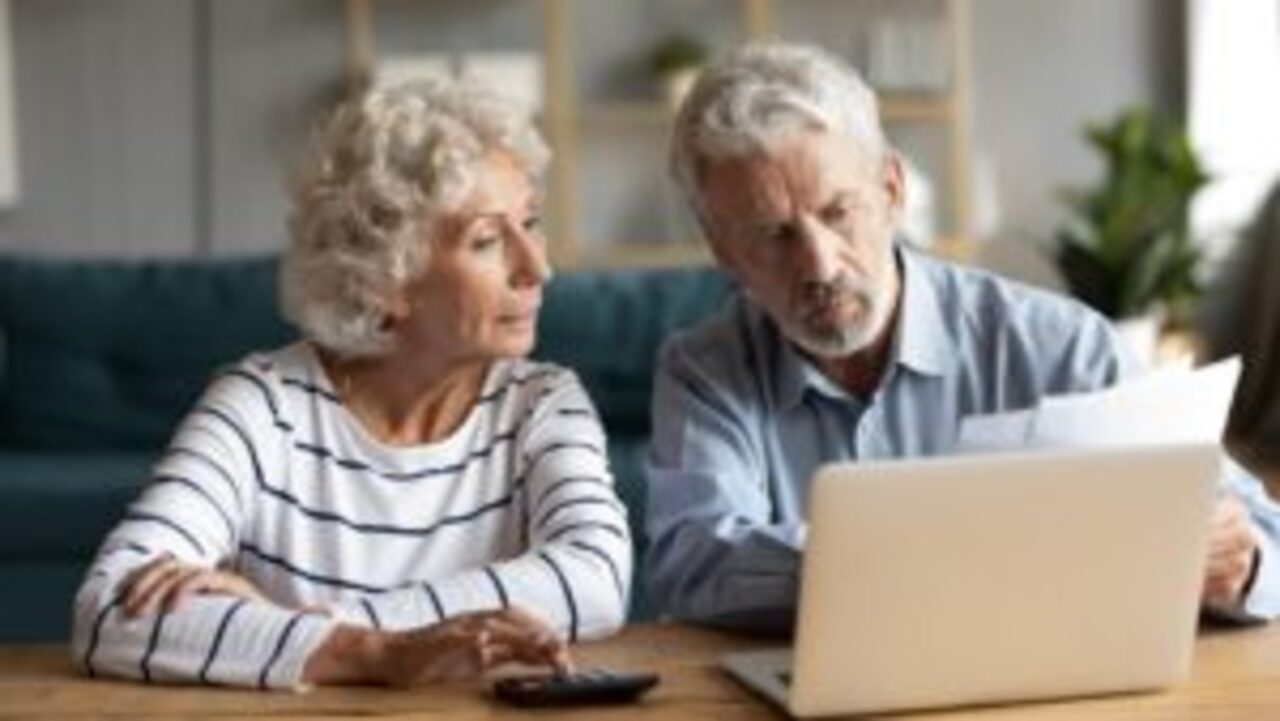 Casal de idosos analisando documento enquanto leem notícia no notebook e usam uma calculadora.