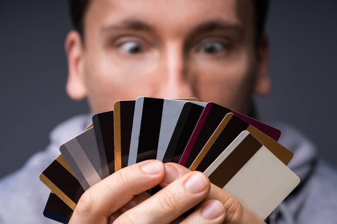 Na imagem, homem desfocado olhando para um leque de cartões de crédito que ele segura em suas mãos. O foco está nos cartões