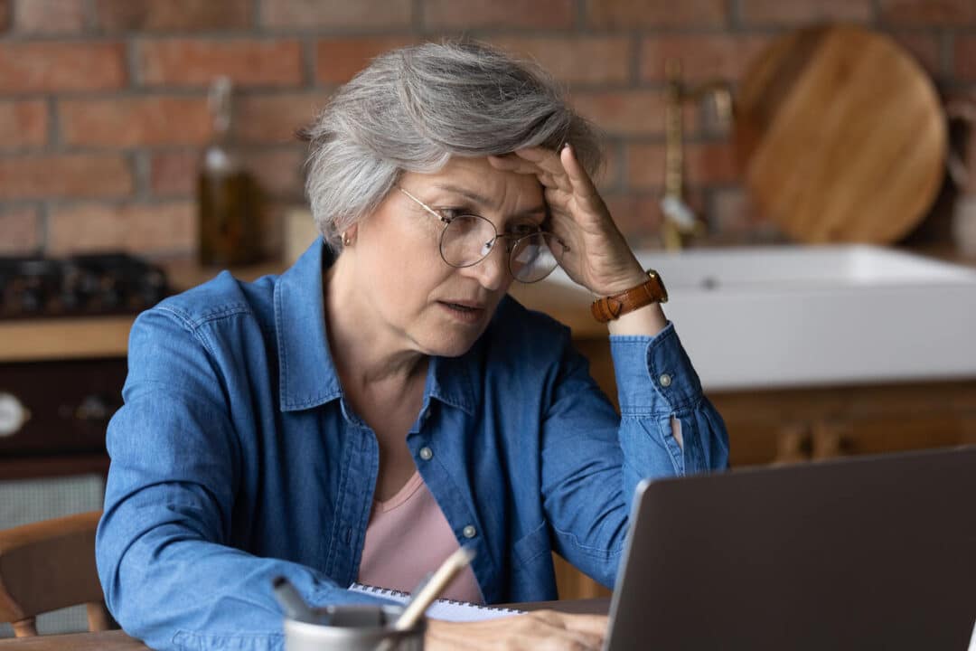 Mulher sentada em frente ao computador com a mão na cabeça e expressão de indignação