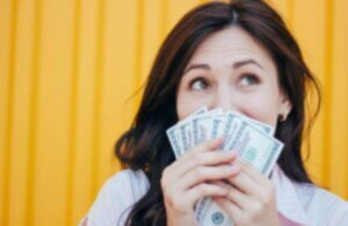 Mulher branca segurando notas de dinheiro em frente a sua boca. Tem um semblante alegre.