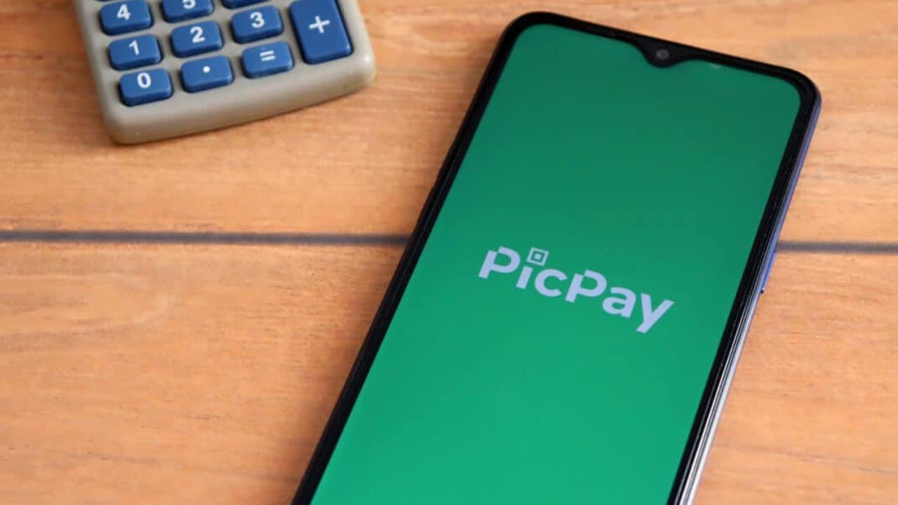 Aplicativo PicPay aberto no celular sobre uma mesa de madeira. À esquerda há um calculadora.