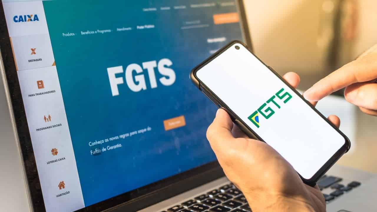 Mão segurando um celular com o logo do FGTS na tela em frente a um notebook que exibe o site da Caixa aberto na página do FGTS