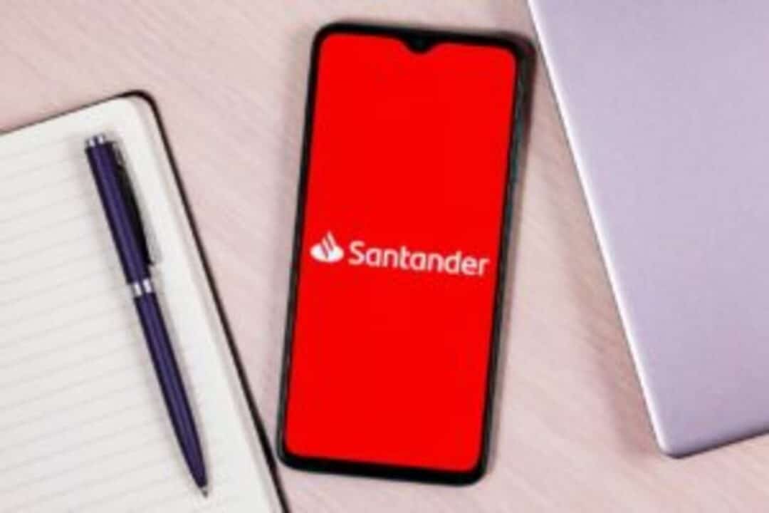 Caderno e caneta de um lado, parte de uma laptop do outro e centralizado na imagem um celular com logo do Santander na tela