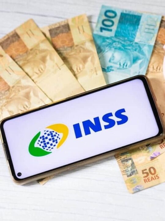 Um celular mostra na tela 'INSS', um cofre de porquinho rosa está ao lado e cédulas de 50 e 100 reais em baixo.