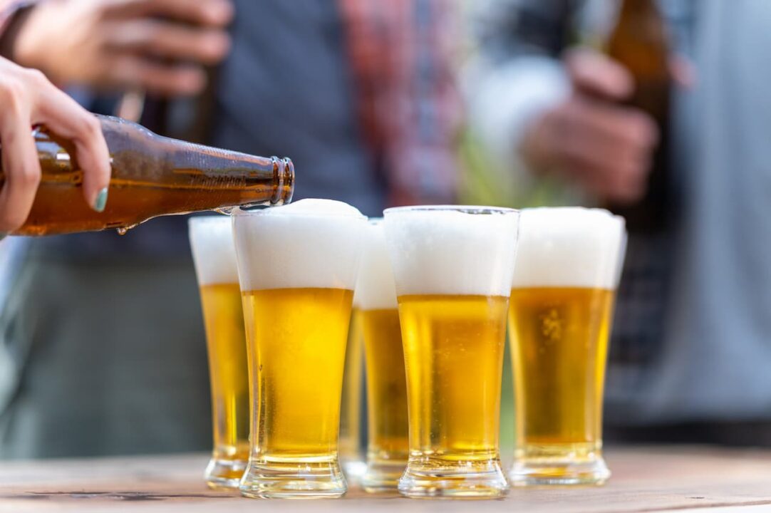 garrafa enchendo copos de cerveja, são vários copos lado a lado. é possível ver pessoas desfocadas ao fundo