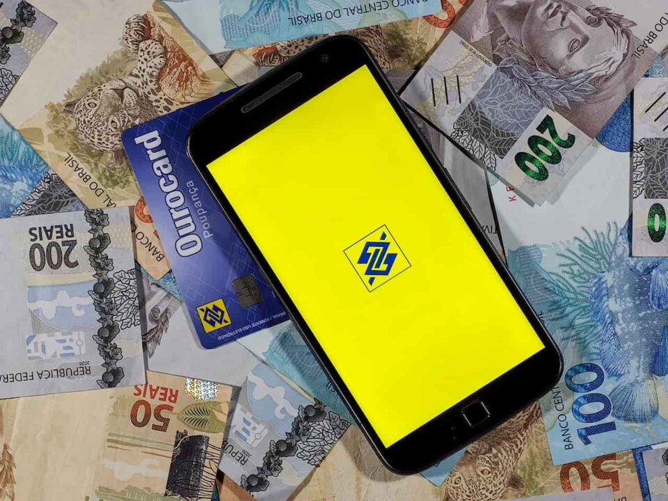 Em cima de notas de 50, 100 e 200 reais e cartão Ourocard, celular exibe logo do Banco do Brasil
