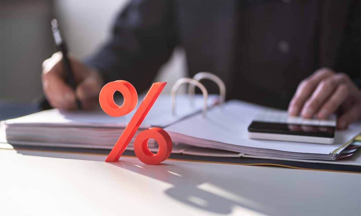 Imagem de um símbolo de porcentagem em frente a um fichario e calculadora representando taxas de juros