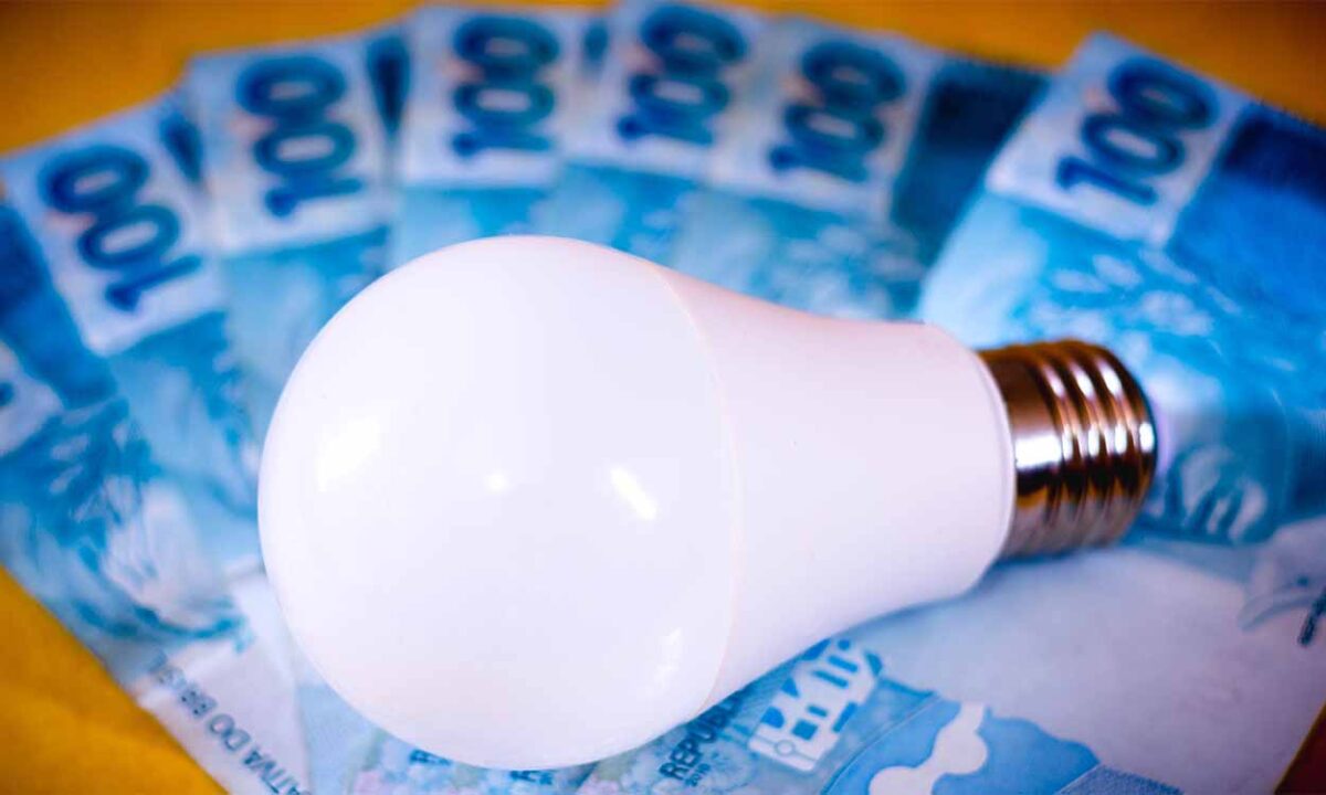 Imagem mostra uma lĂąmpada em cima de vĂĄrias notas de cem reais.