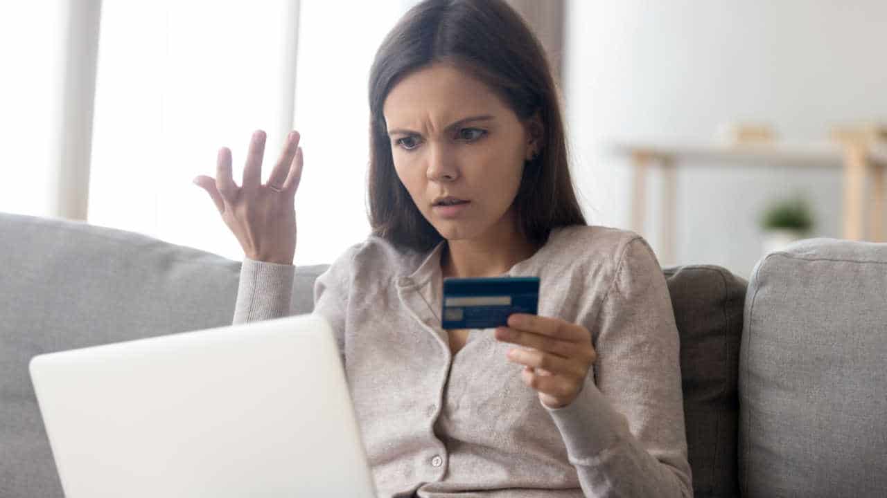 mulher com expressão assustada e indignada olhando para o notebook, com cartão de crédito na mão