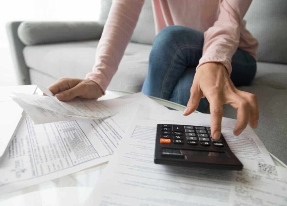 Mulher usando calculadora para calcular dívidas a pagar. A sua frente há uma mesa cheia de papeis.