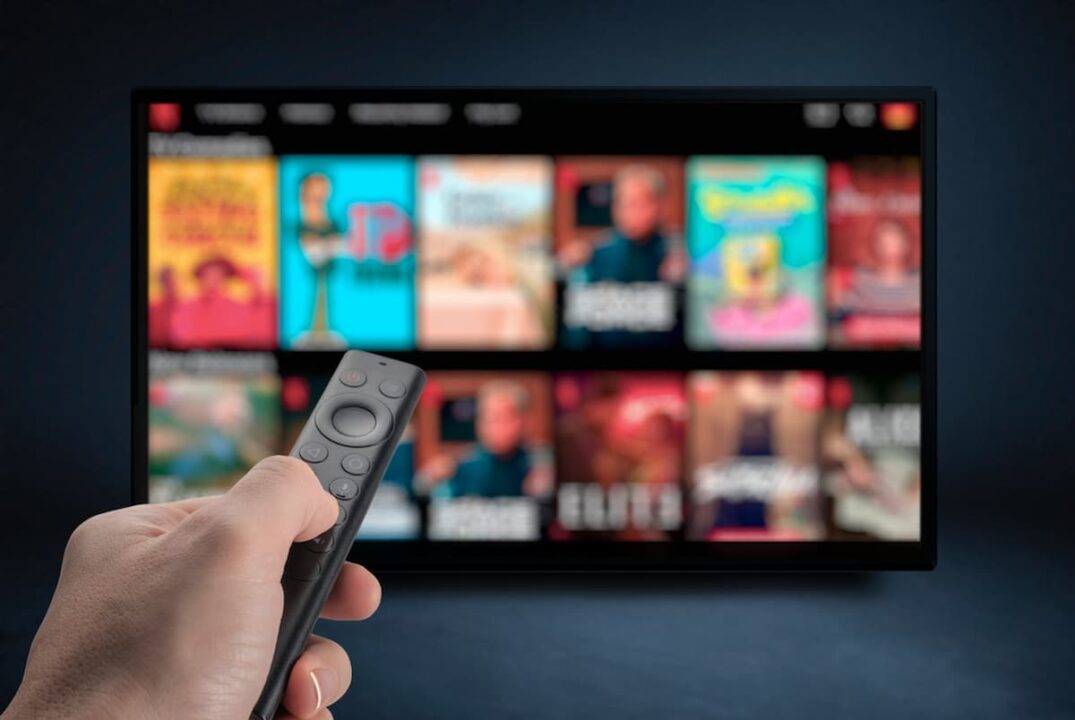 Pessoa apontando controle para televisão, que está mostrando alguma plataforma de serviço de streamings.
