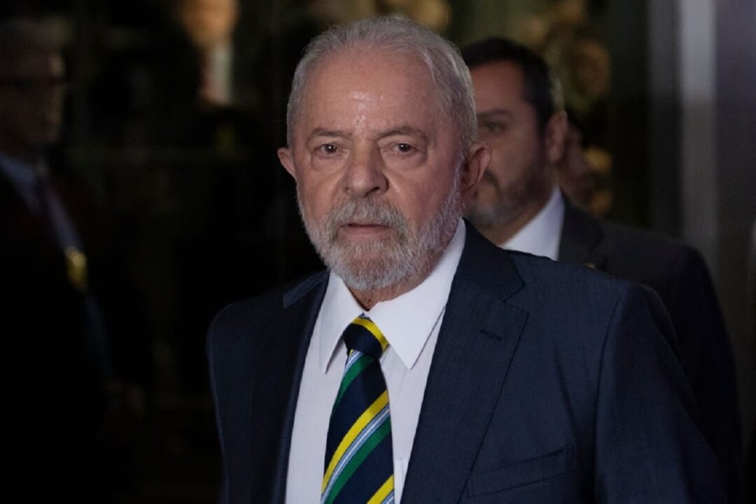 Presidente Lula com expressão séria. Ao fundo, há dois homens desfocados, um quase invisível