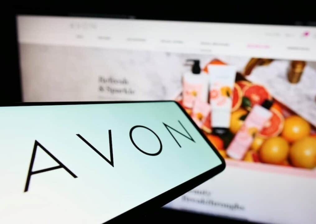 Na imagem, celular com o logo da Avon na tela e atrás um computador com o site aberto.