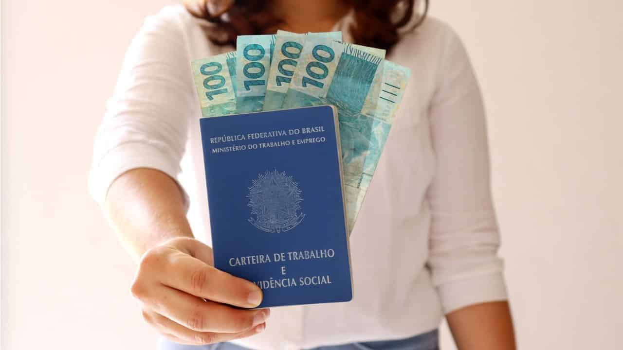 Na imagem, mulher segurando uma carteira de trabalho com notas de 100 reais dentro