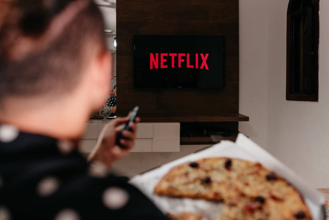 Homem comendo pizza e segurando controle remoto da televisão na mão. Ao fundo, uma TV exibindo o logotipo da Netflix.