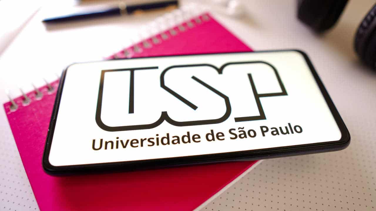 Tablet em cima de um caderno vermelho. Ele exibe o logo da USP. Ao lado do ccaderno, uma caneta