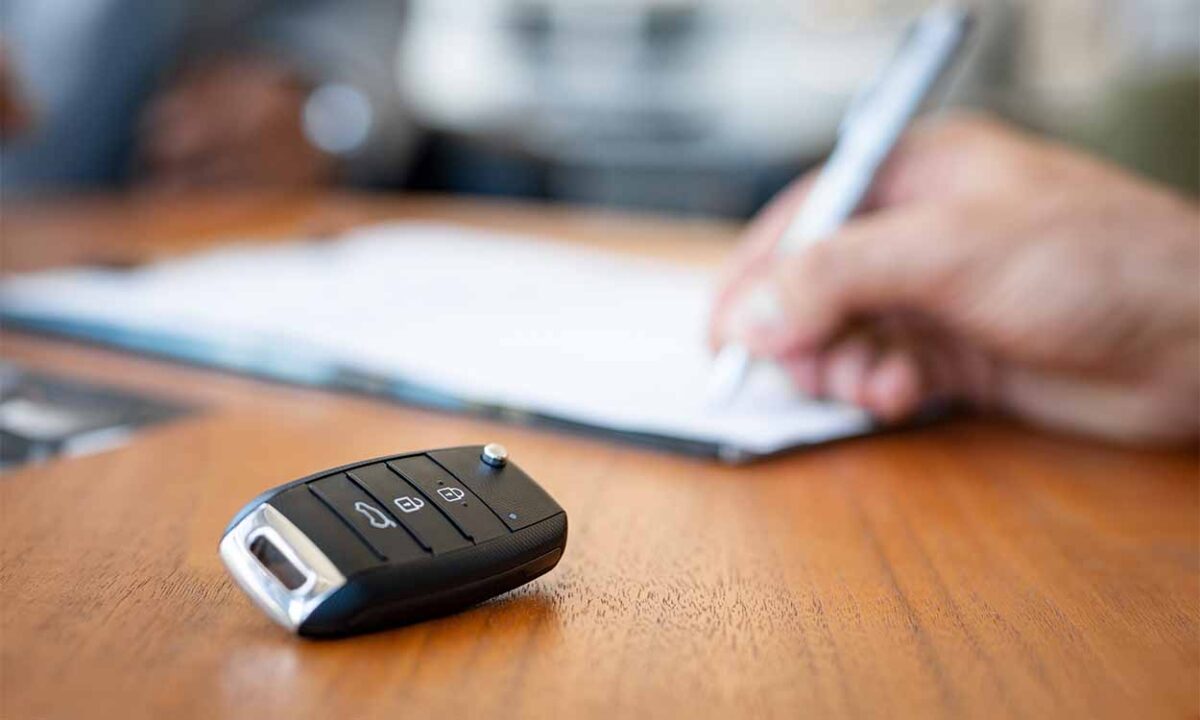 Chave do carro em cima da mesa e ao fundo, mão masculina assinando contrato de aluguel.