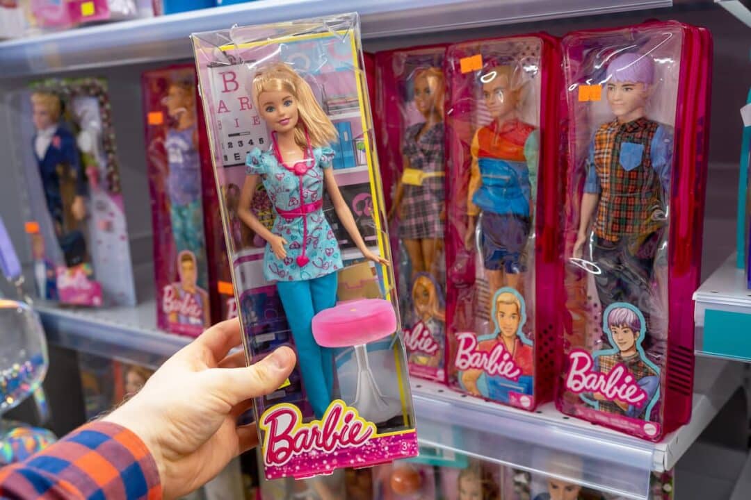 Mão segurando uma embalagem com uma Barbie dentro e bonecas da Barbie ao fundo, numa prateleira