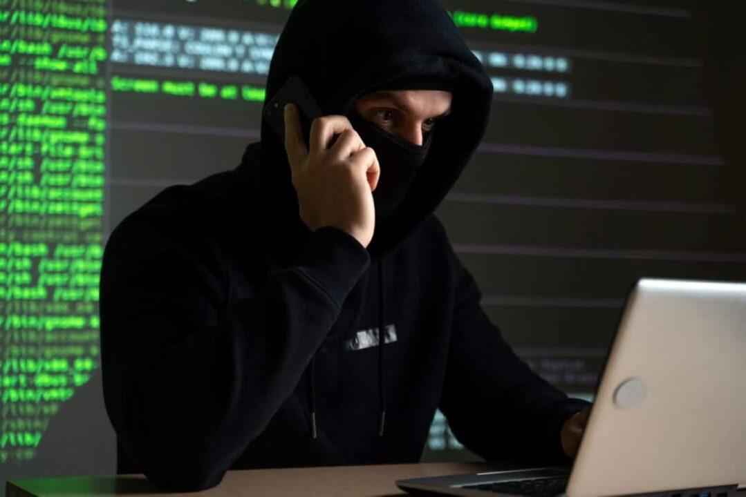 Cibercriminoso com parte do rosto coberta com capuz e máscara usando o computador para aplicar golpes, por trás dele há uma tela com códigos de programação.