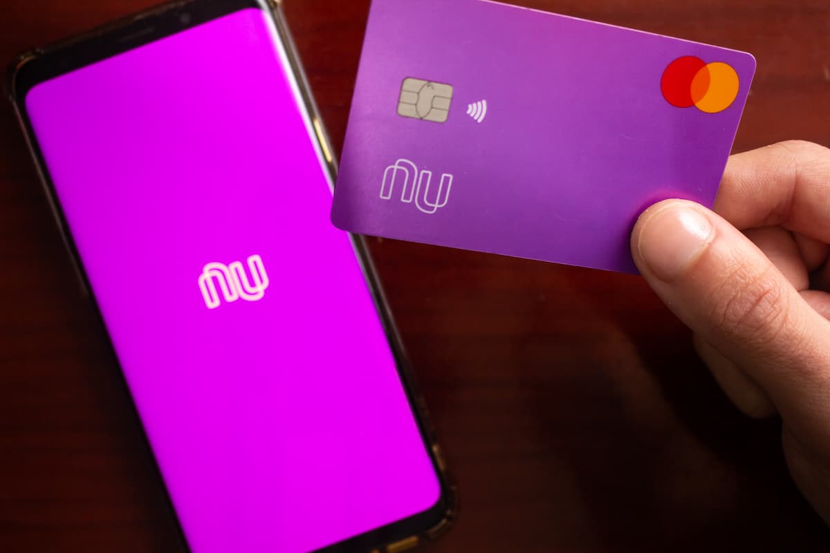 A imagem mostra uma mão segurando um cartão de crédito Nubank e um celular com a logo do mesmo.