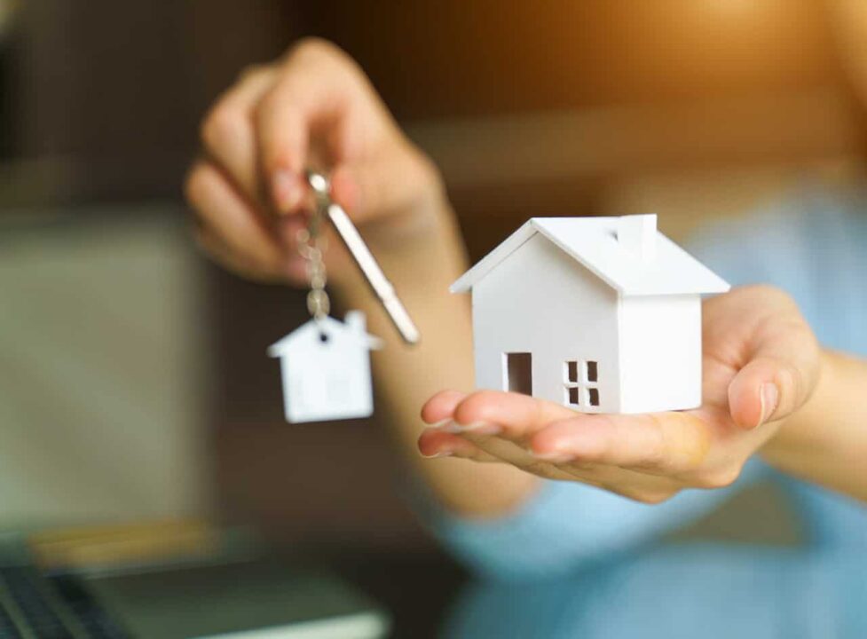 Pessoa segurando miniatura de uma casa branca e chaves com chaveiro em formato de uma casinha.