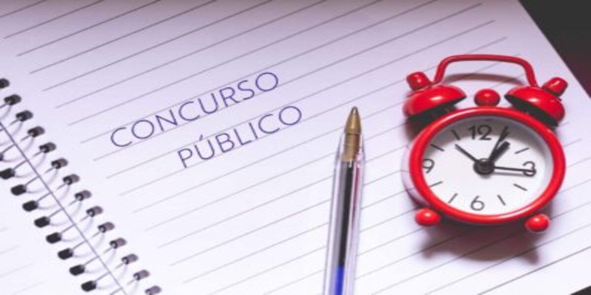 Um caderno com os dizeres "Concurso Público", uma caneta e um relógio vermelho em cima