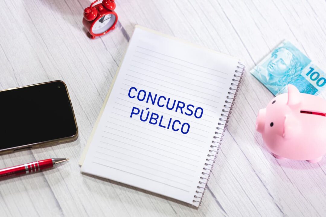 caderno concurso público, celular, relógio, porquinho e dinheiro