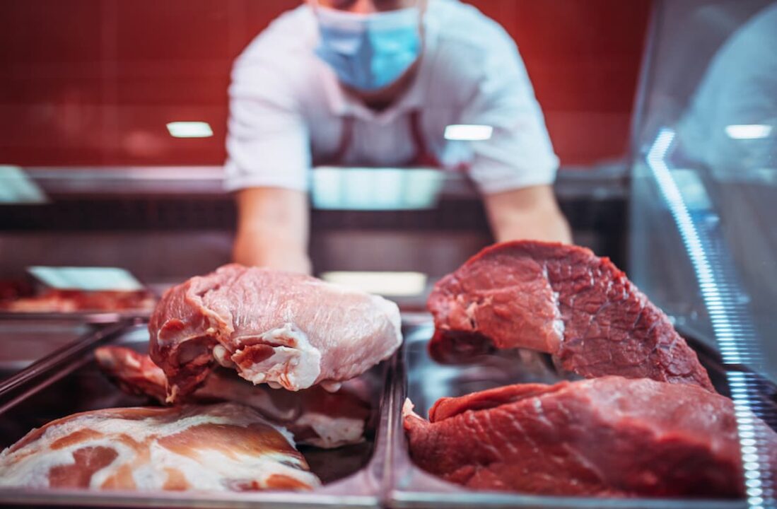Carne crua exposta em vitrine de açougue, com homem de máscara ao fundo.
