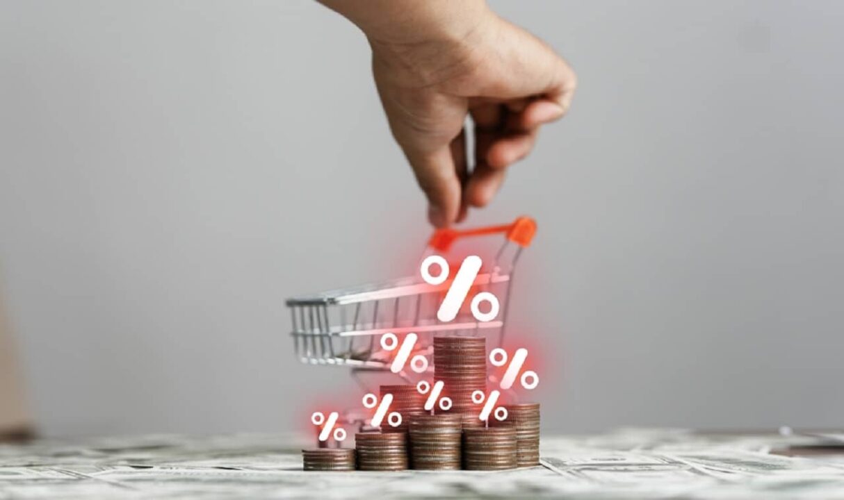 Imagem de um carrinho de mercado em miniatura e moedas com o símbolo "%" em cima, simbolizando índices como inflação e deflação
