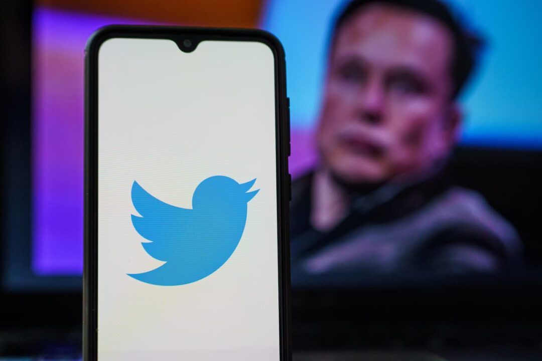 celular com logo do Twitter, ao fundo uma imagem desfocada de Elon Musk