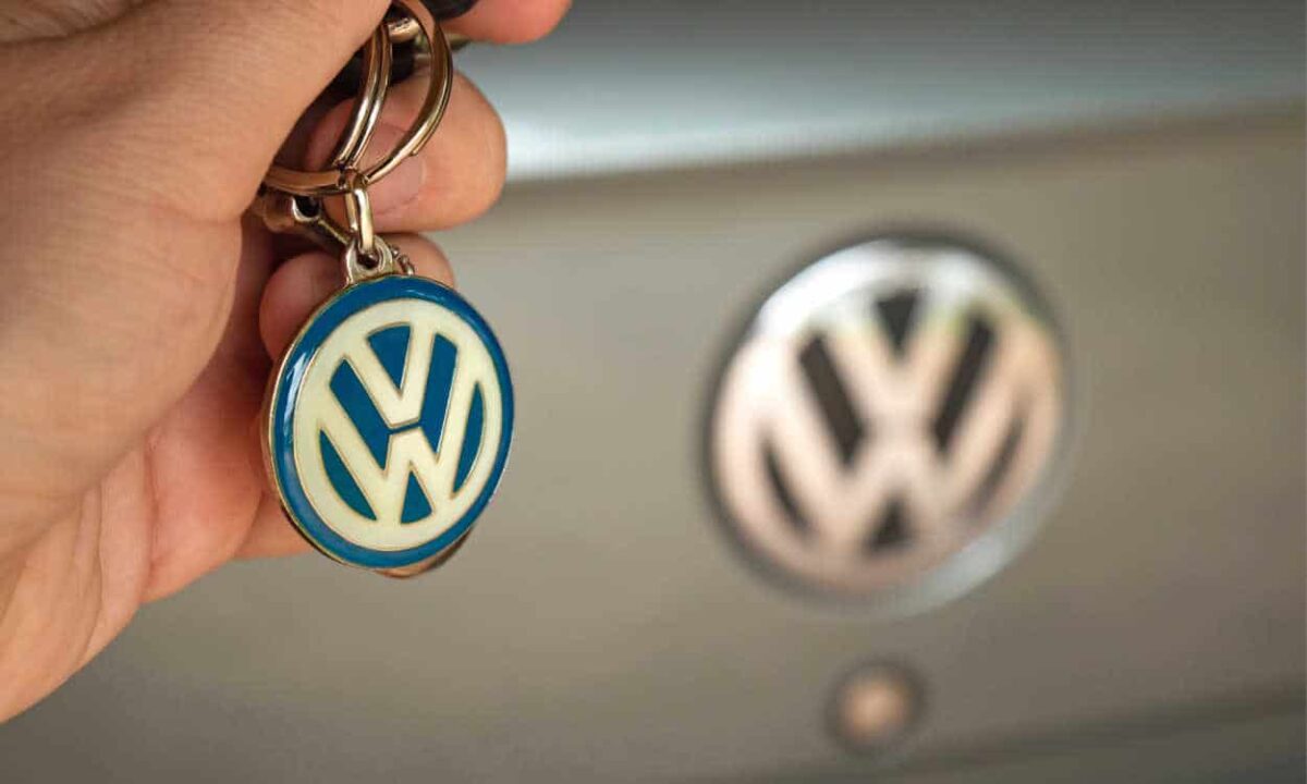 Mão segurando um chaveiro da Volkswagen com a traseira de um carro da marca ao fundo