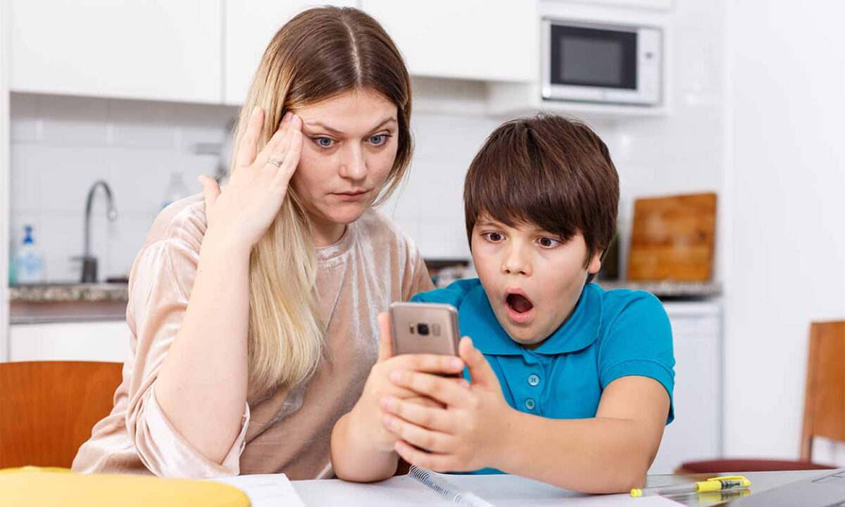 Mulher - com expressão estressada - sentada ao lado de uma criança segurando um celular, que se mostra surpresa