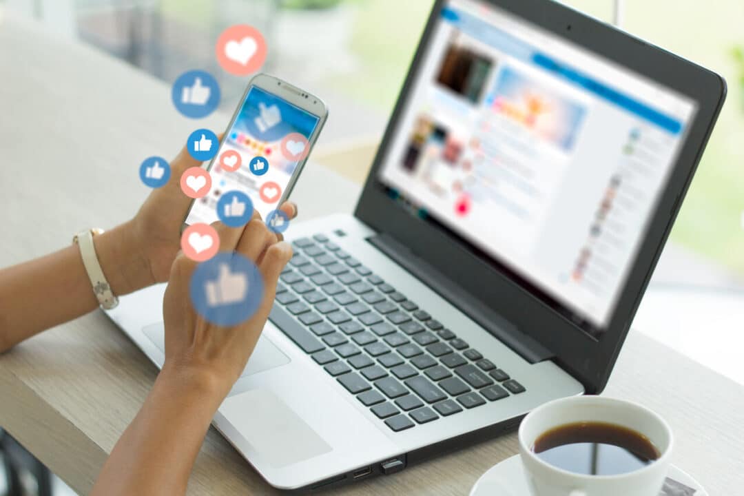 Na imagem, pessoa usando o notebook e o celular com vários símbolos de likes e curtidas em redes sociais.