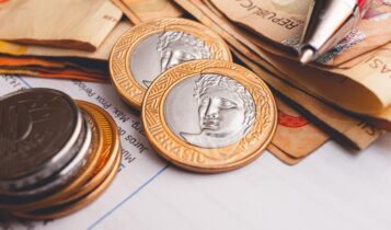 Imagem de moedas, cédulas de real e uma caneta em cima de um papel.