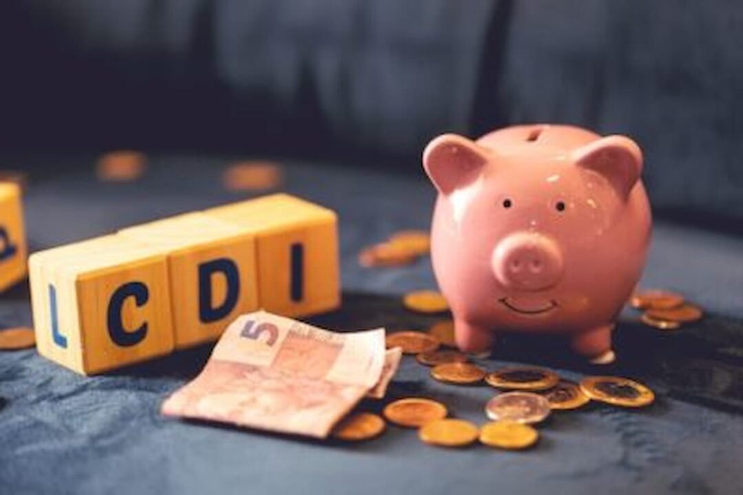 Fundo cinza. À frente, blocos de madeira formando a sigla CDI, uma cédula de 5 reais ao lado, moedas e um cofre em forma de porquinho.