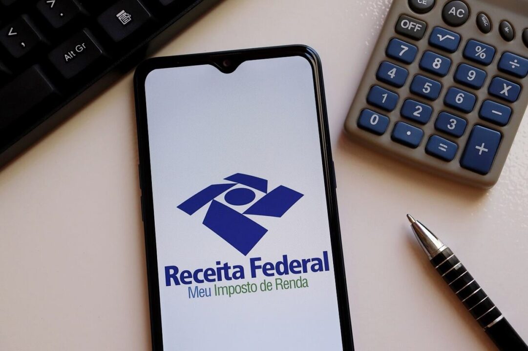 Na imagem, um celular com a logo da Receita Federal na tela, uma calculadora, uma caneta e parte do teclado de um computador