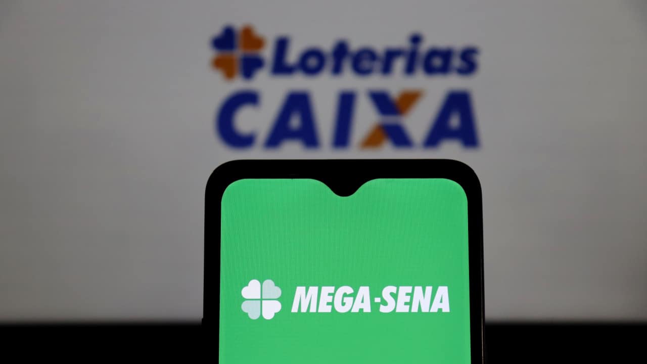 Celular mostrando logo da Mega-Sena. Ao fundo, logo das Loterias Caixa aparece desfocado.