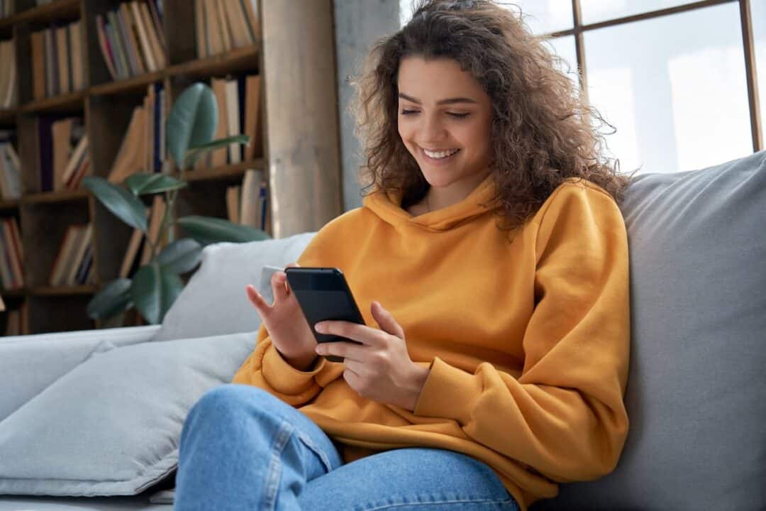mulher sentada em um sofá, usando um celular, com a expressão feliz.