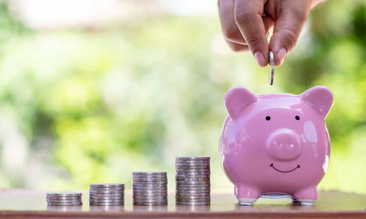 Pessoa colocando uma moeda dentro de um porquinho que está ao lado de várias pilhas de moedas