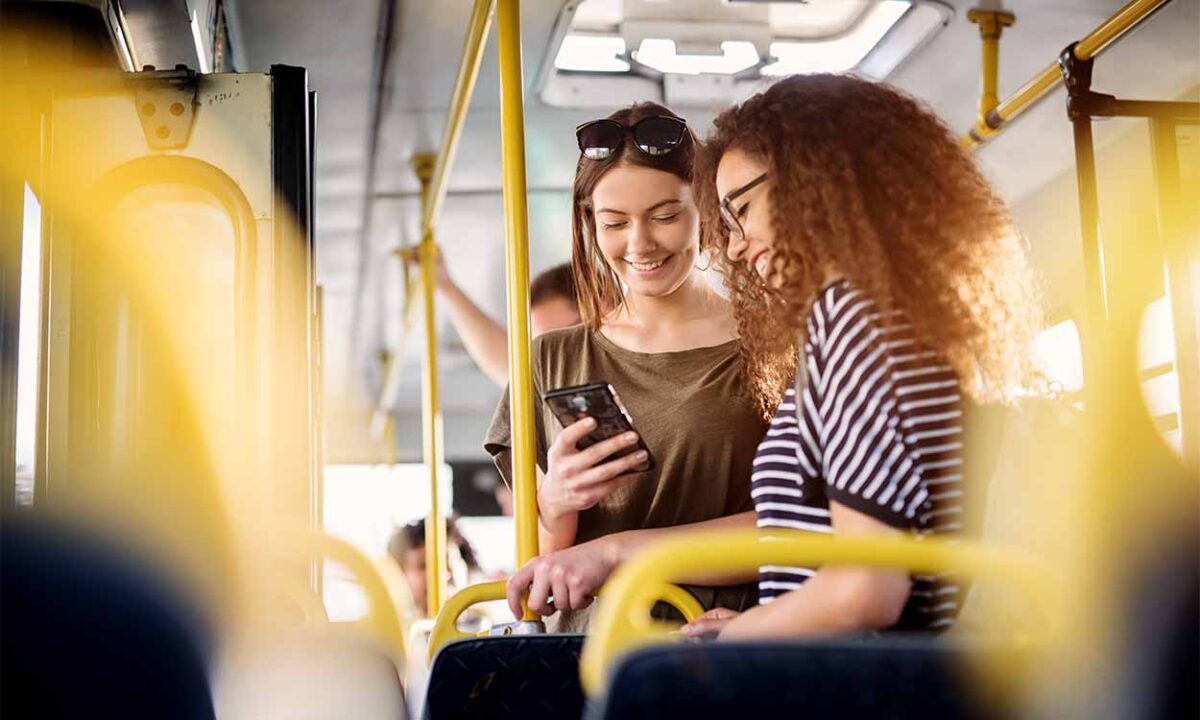 Duas jovens em pé em um ônibus. Elas sorriem e leem notícia em um celular.