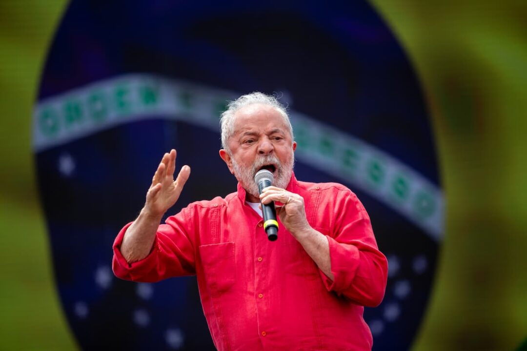 Presidente Lula falando no microfone, com camisa vermelha e bandeira do Brasil ao fundo.