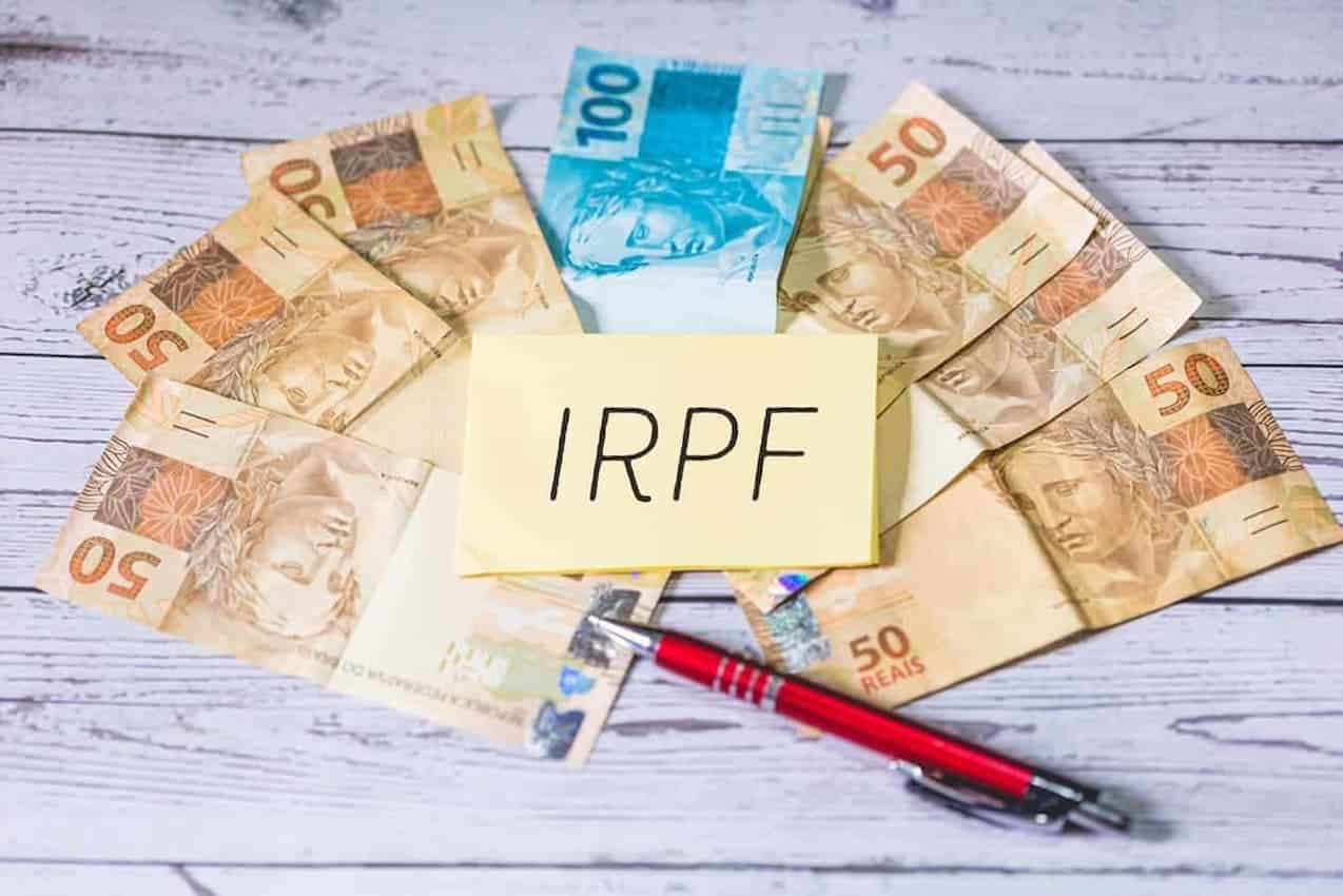 Papel com "IRPF" escrito e notas de dinheiro e caneta ao redor.